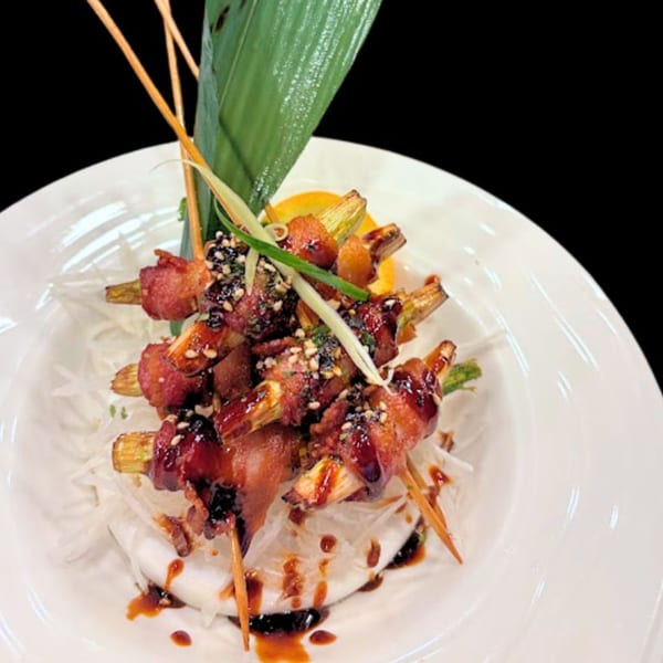 Bacon-wrapped asparagus skewers with sauce and sesame seeds, garnished with a green leaf on a white plate.