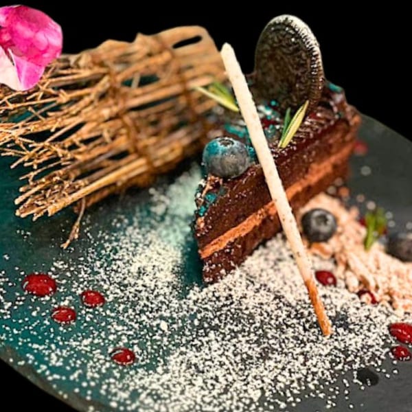 A slice of chocolate cake with blueberries, an Oreo, and powdered sugar on a dark plate.