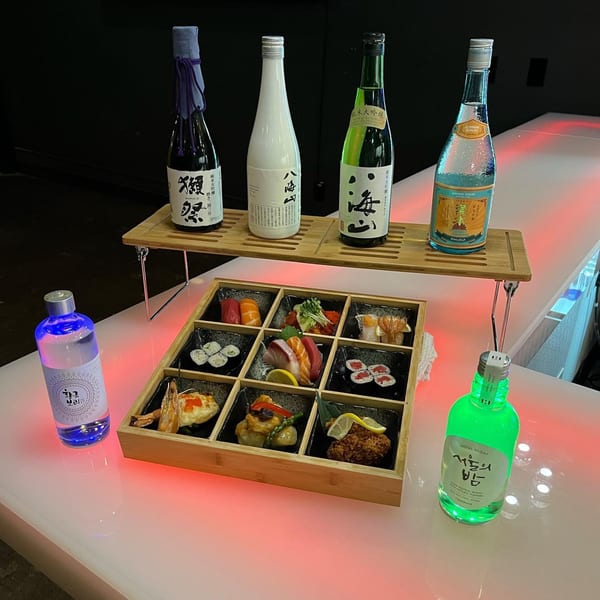 A bento box of Sushi Hiro's sushi and sashimi, with various sake bottles on a glowing red bar.