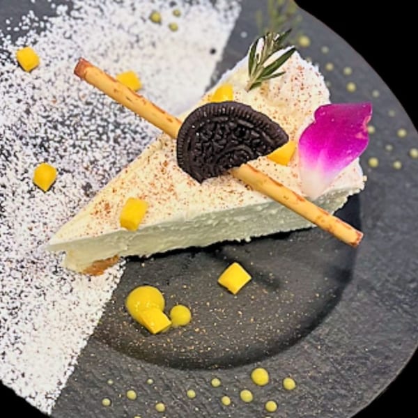 A slice of cheesecake with an Oreo, mango, rosemary, and a pink petal on a dark plate.